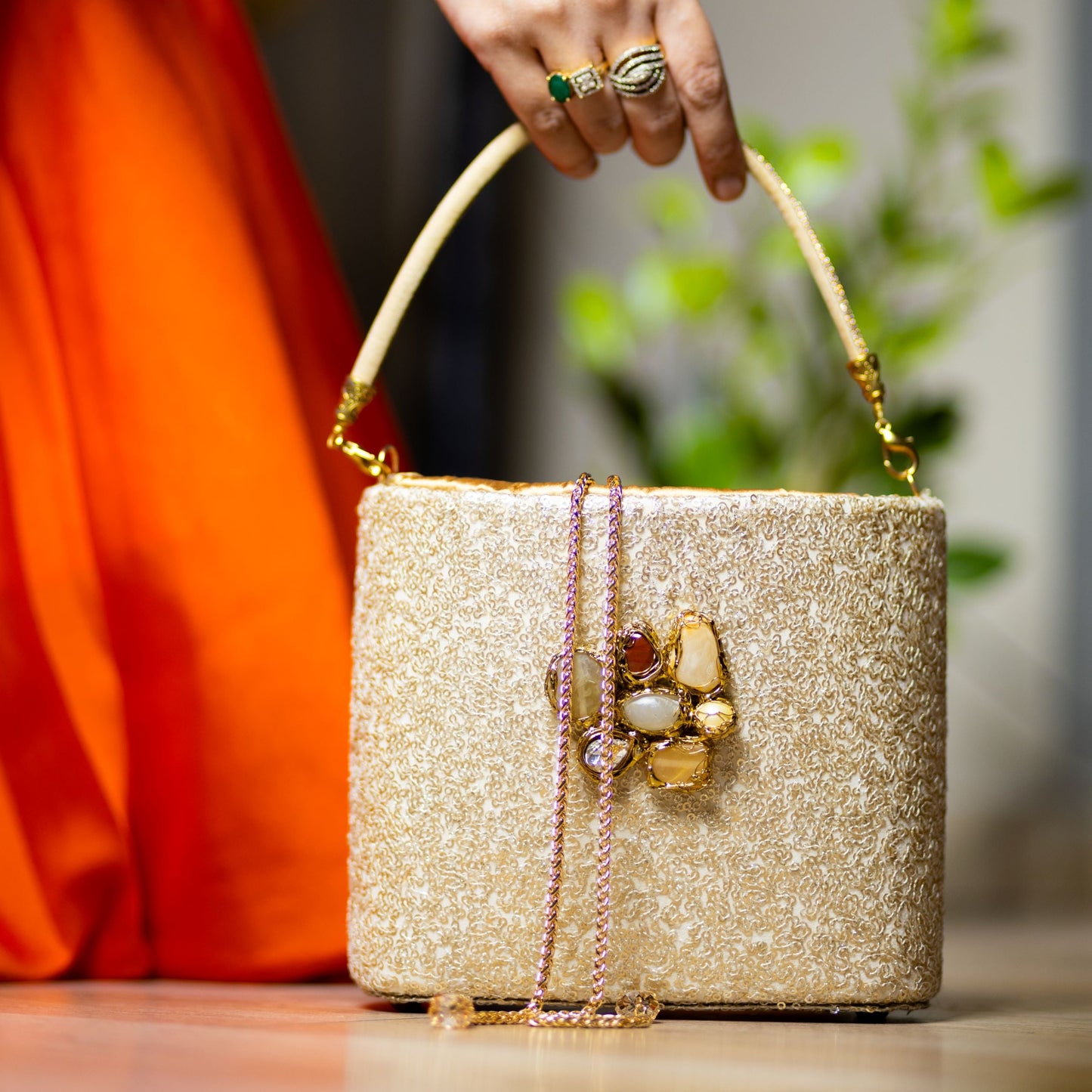 A gold sequin clutch with crystal embellishments and a decorative handle being held by a person with a orange garment in the background.