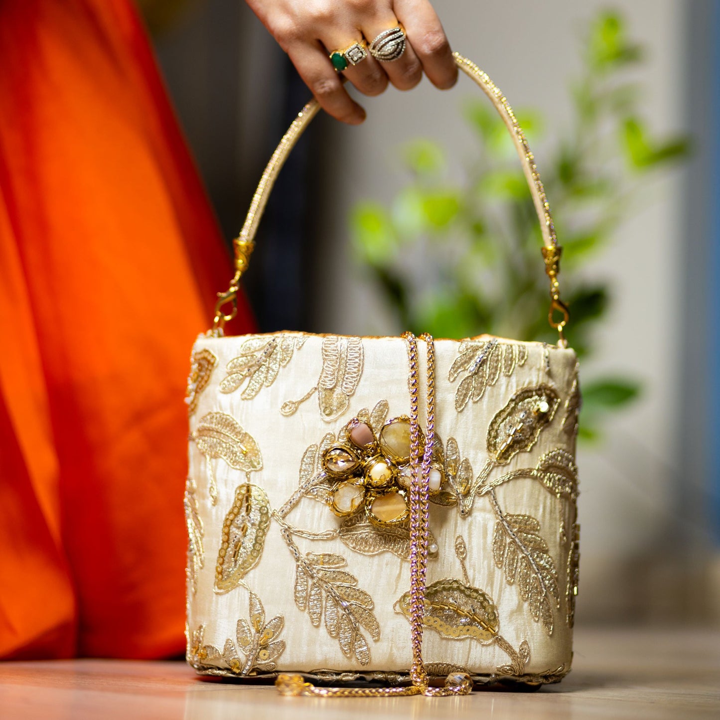 An ivory clutch with floral gold work and pearl embellishments, held by a person with a orange garment in the background.
